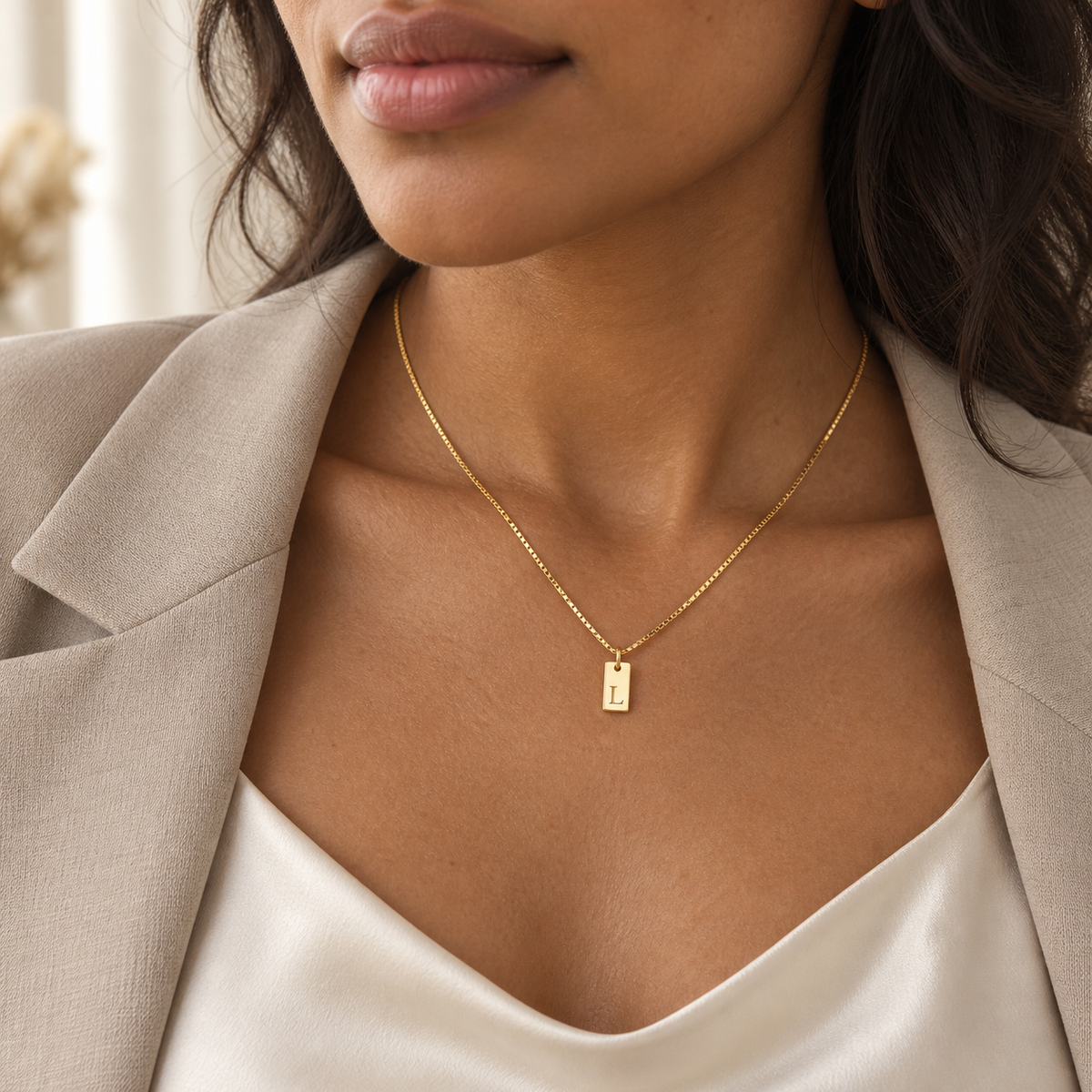 Woman wearing a petite gold initial tag necklace with an ivory satin top and beige blazer