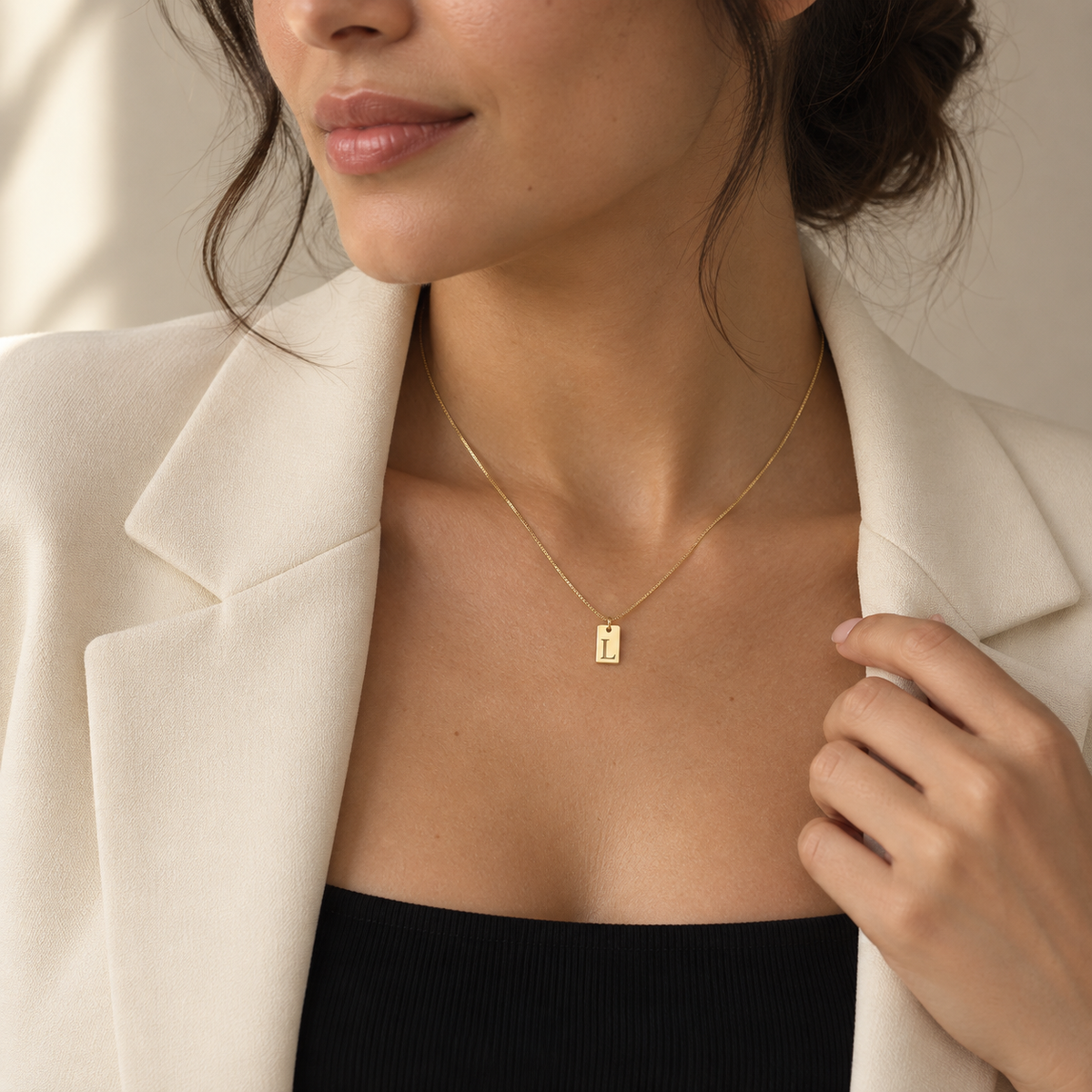 Woman wearing a dainty gold letter L necklace with a black top and cream blazer