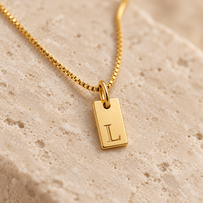 Close-up of a polished gold initial tag pendant with engraved letter L on an ivory stone surface